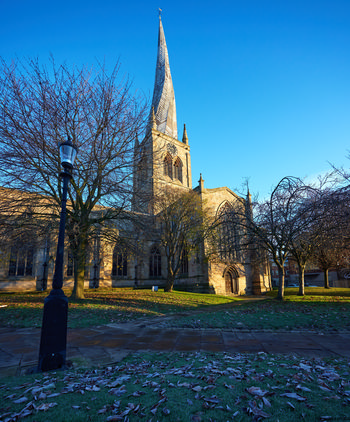 Church of St Mary and All Saints winter frost The photograph presents a landscape view of the Church of St Mary and All Saints in Chesterfield during a winter morning. The image prominently features the church, known for its distinctive twisted and leaning spire, which rises above the surrounding trees. A light winter frost is visible covering the ground and fallen leaves, indicative of the cold season. The sunlight casts long shadows across the church grounds, illuminating the stone architecture and the leafless branches of nearby trees. The photograph captures the architectural details of the Church of St Mary and All Saints, with its Gothic elements and famous crooked spire, under clear blue winter skies.